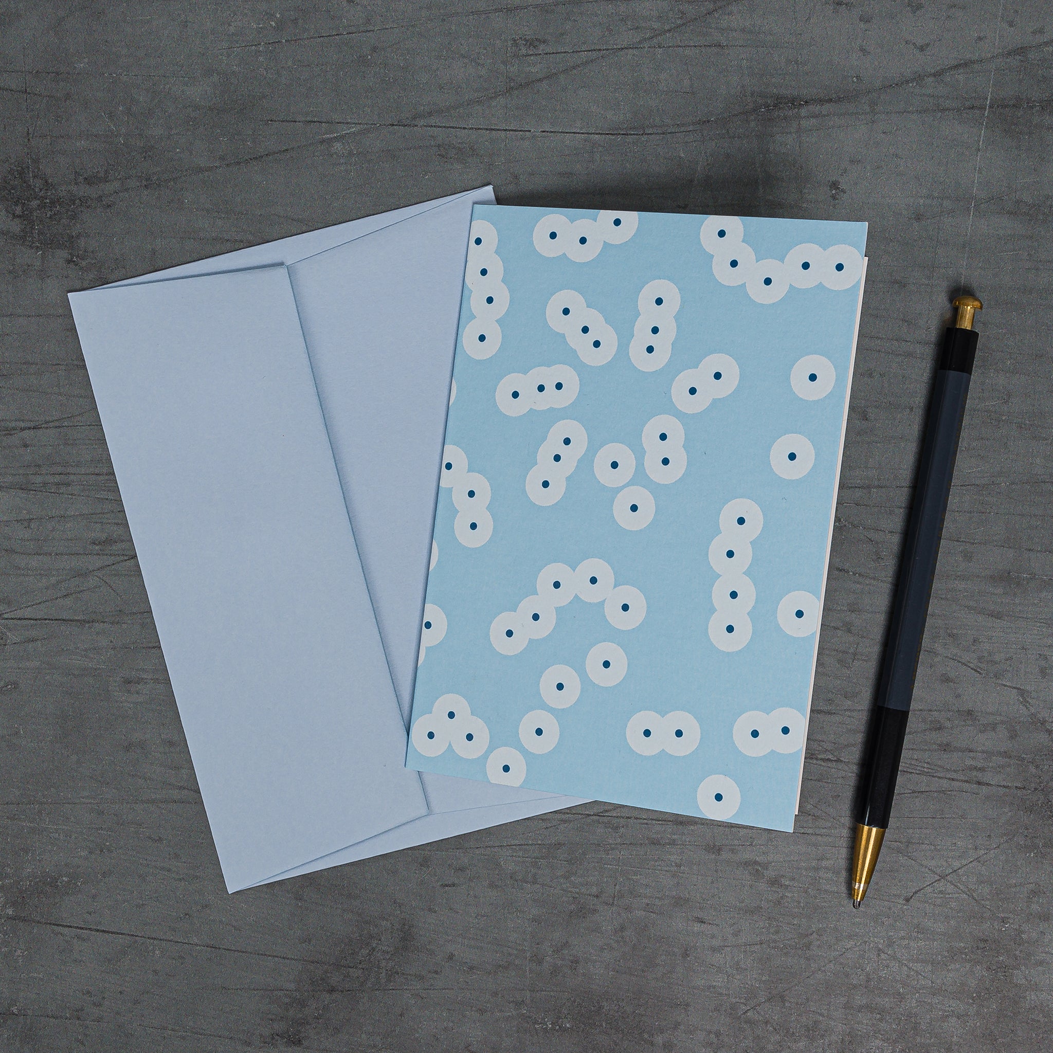 Aerial view, slanted, of Stack (blue) greeting card and matching pale blue envelope. Pattern: schematic version of frog spawn, formed of clustering circle motifs