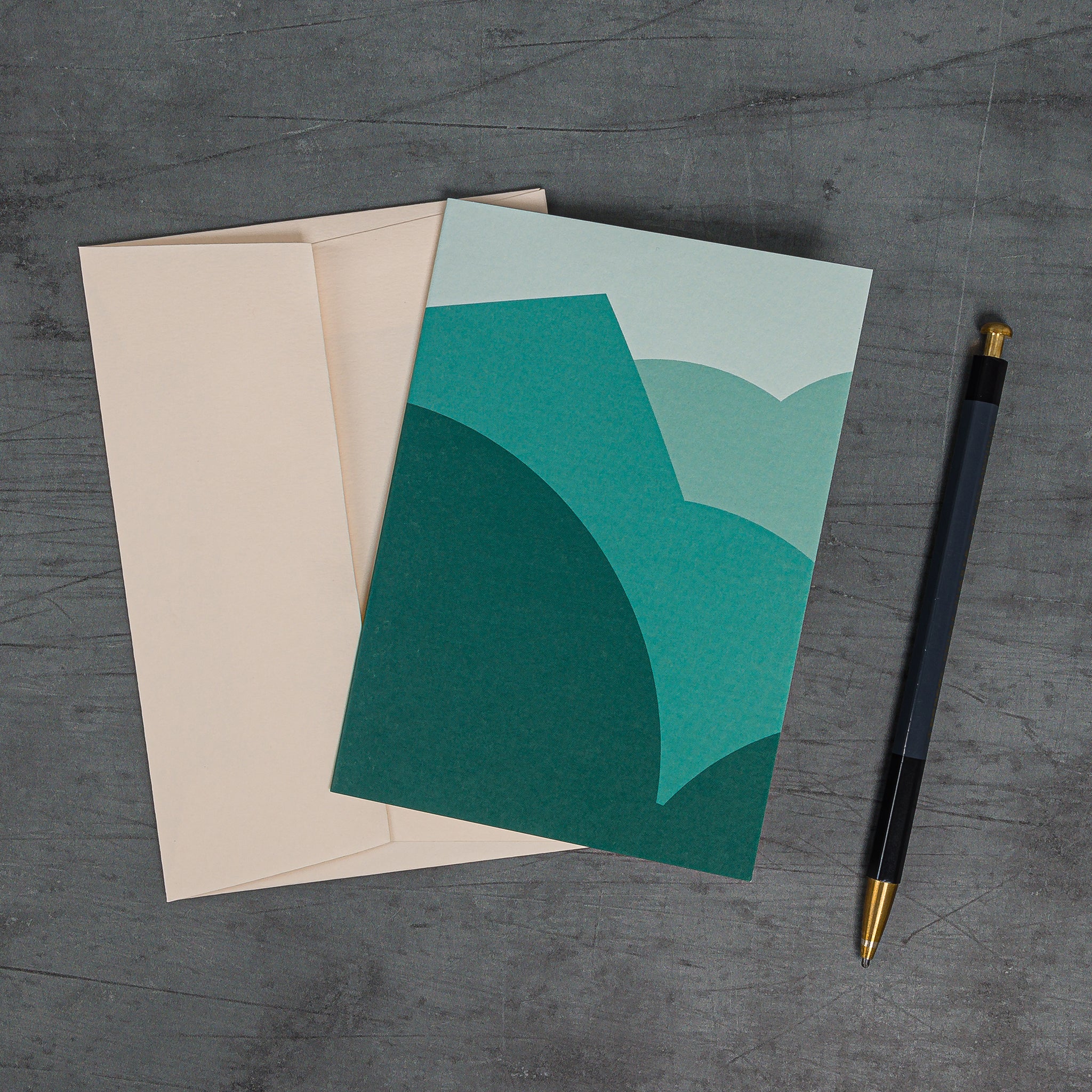 Aerial view, slanted, of the Stack (teal) greeting card. Layerd curves and angles resembling a schematised landscape. Teal colour scheme and coordinating peach-coloured envelope