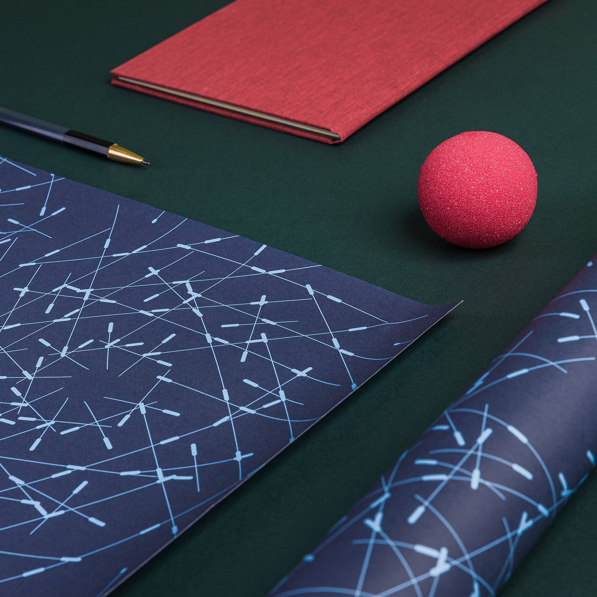 Aerial view of two sheets of Briar wrapping paper - one flat, one folded. Schematised pattern of overlapping dashed lines, radially arranged, inspired by thickets of shrubs in nature. Dark blue background, pale blue lines