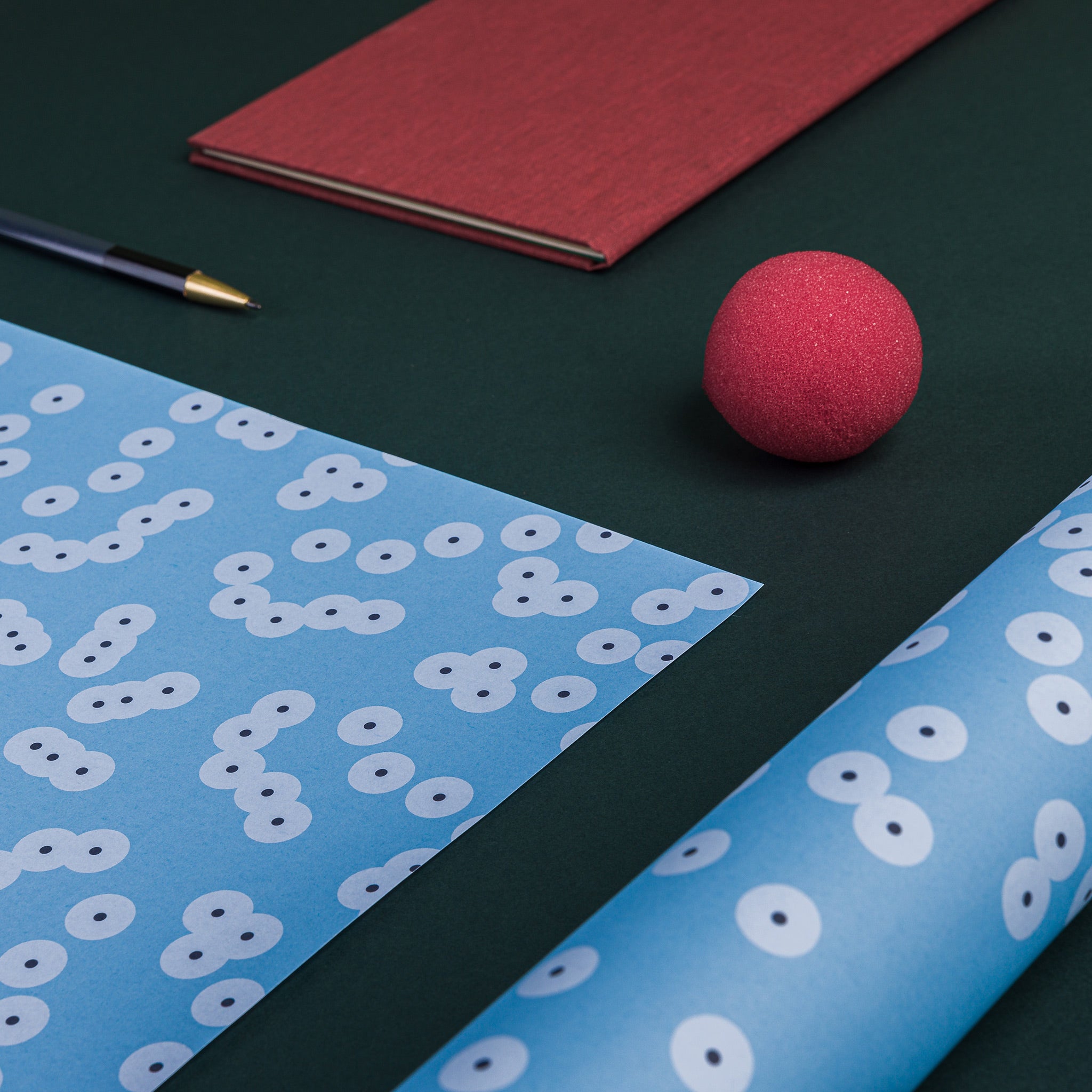 Angled view of Spawn (blue) wrapping paper, showing part of one flat sheet and one rolled sheet. Pattern: clusters of circles resembling frog spawn, pale blue colour scheme