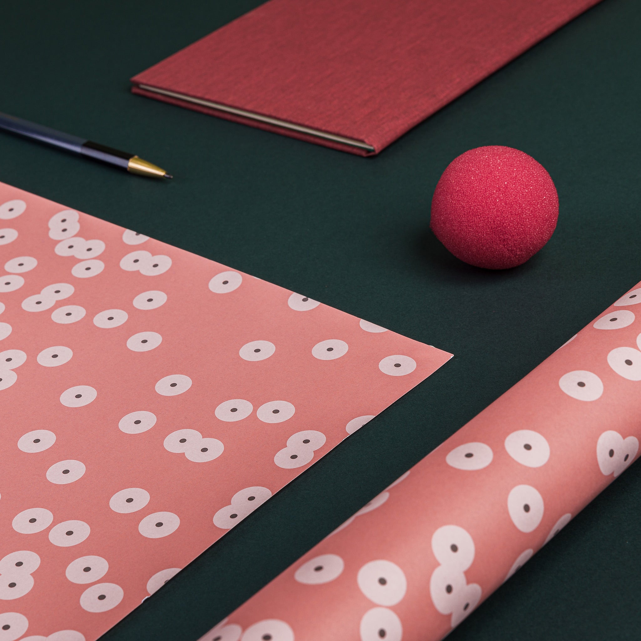 Slanting view of two sheets of Spawn (orange) wrapping paper - one flat, one rolled. Design: schematic version of frog spawn