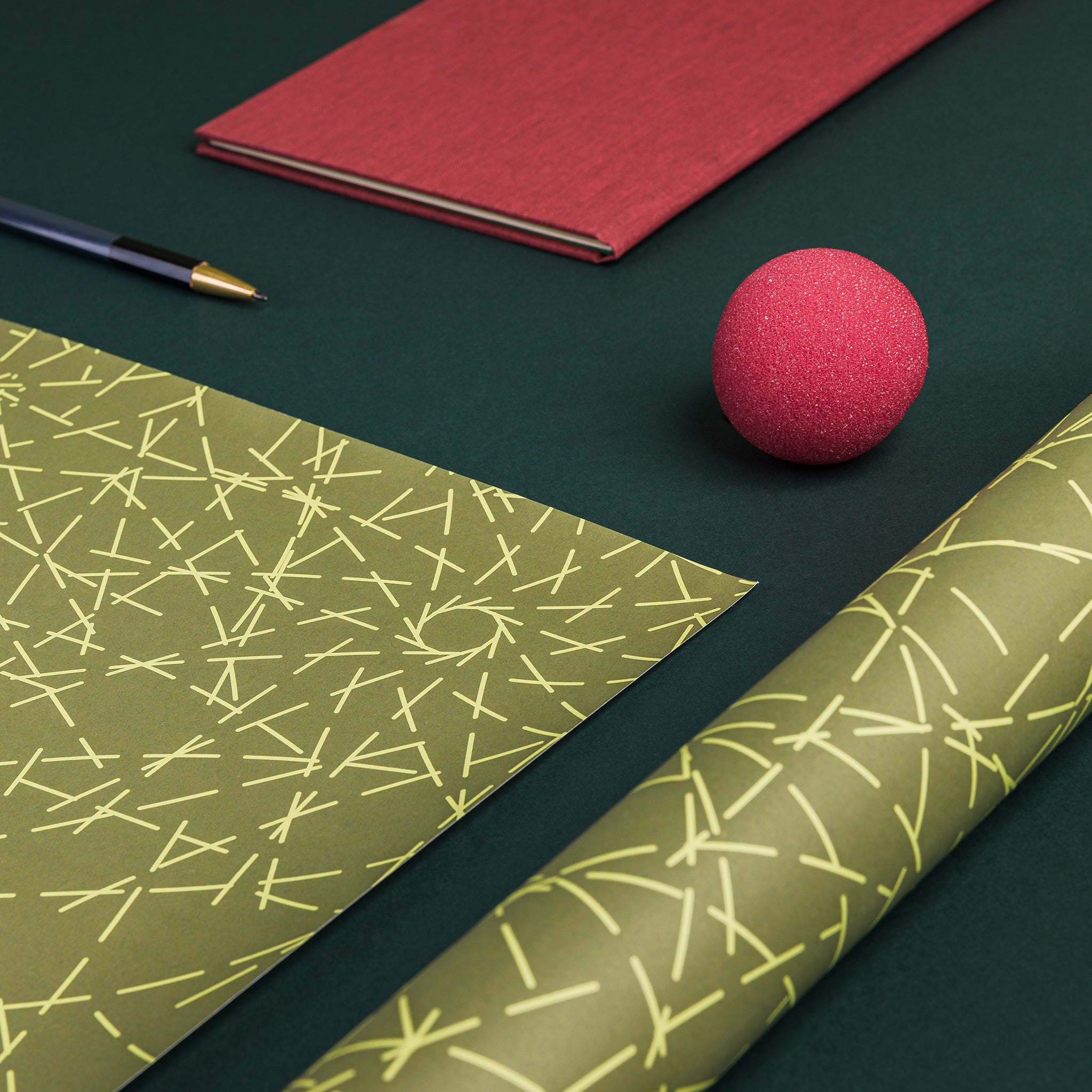 Partial view of two sheets of Turf gift wrap - one flat, one folded. Mid-green background with pale yellowish green, dashed lines. Irregular pattern of overlapping, radiating lines inspired by grass plants in a flower meadow
